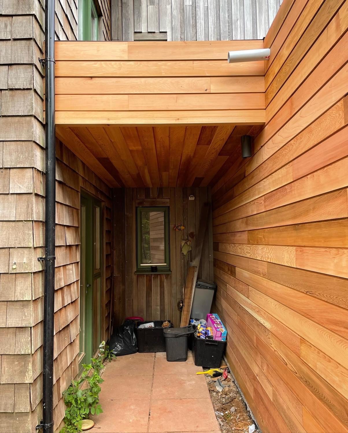 A small, partially covered outdoor space surrounded by wooden walls with a mix of vertical and horizontal planks. It includes various items like black containers, a trash bag, a toy box, and some board-like materials leaning against the wall.