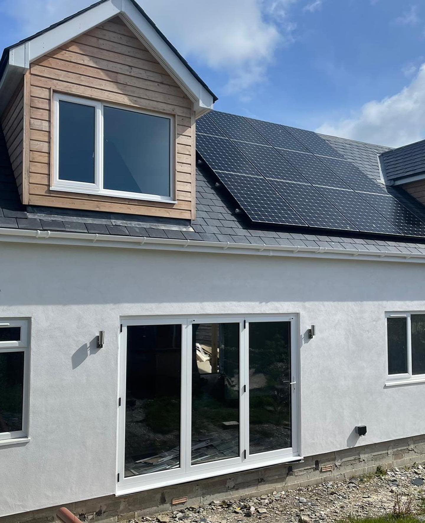 A white house with a dormer window, solar panels on the roof, and large glass patio doors. The ground outside appears unfinished, with some gravel and patches of grass. The sky is partly cloudy.