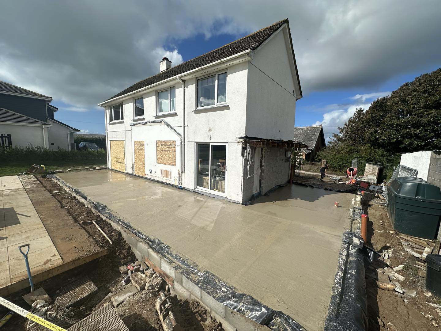 A two-story white house undergoing renovation, with a newly poured concrete foundation extending from the back. Plywood covers some windows, and construction materials are scattered around. A cloudy sky is overhead.