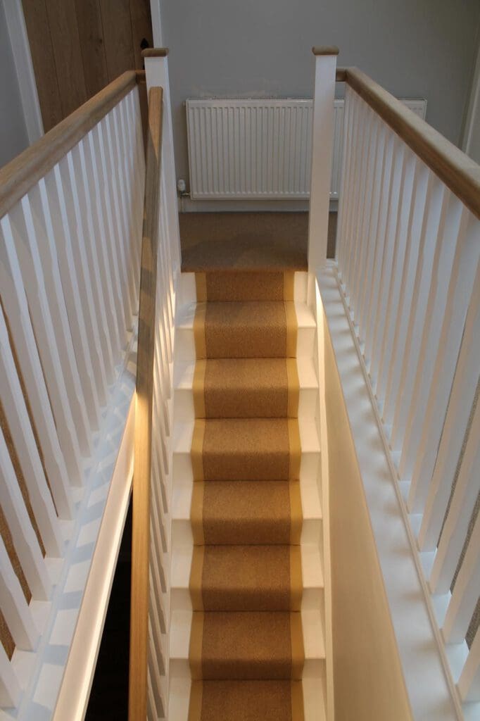 A staircase with beige carpet runner bordered by white. Wooden handrails and white spindles line the sides. A white radiator is visible at the top of the stairs. The stairs are illuminated by soft lighting.
