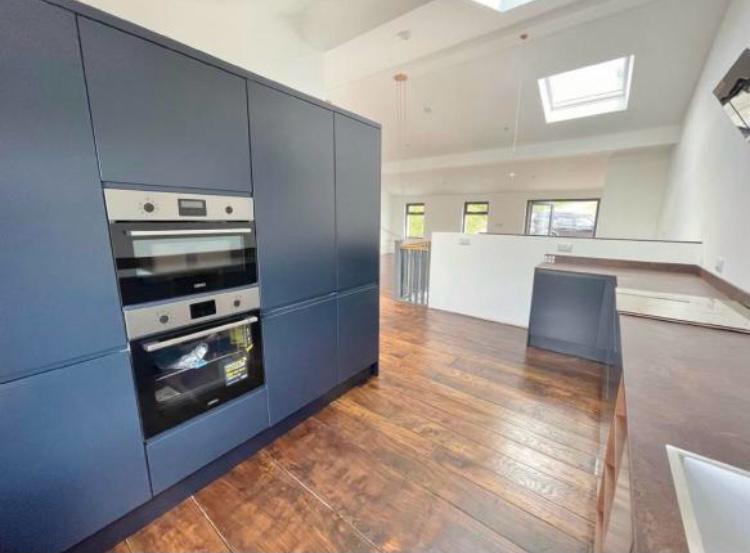 Modern kitchen with dark blue cabinets, built-in stainless steel oven, and wooden floor. Bright space with skylights and multiple windows. Countertops are clean and uncluttered. Minimalist design with ample natural light.