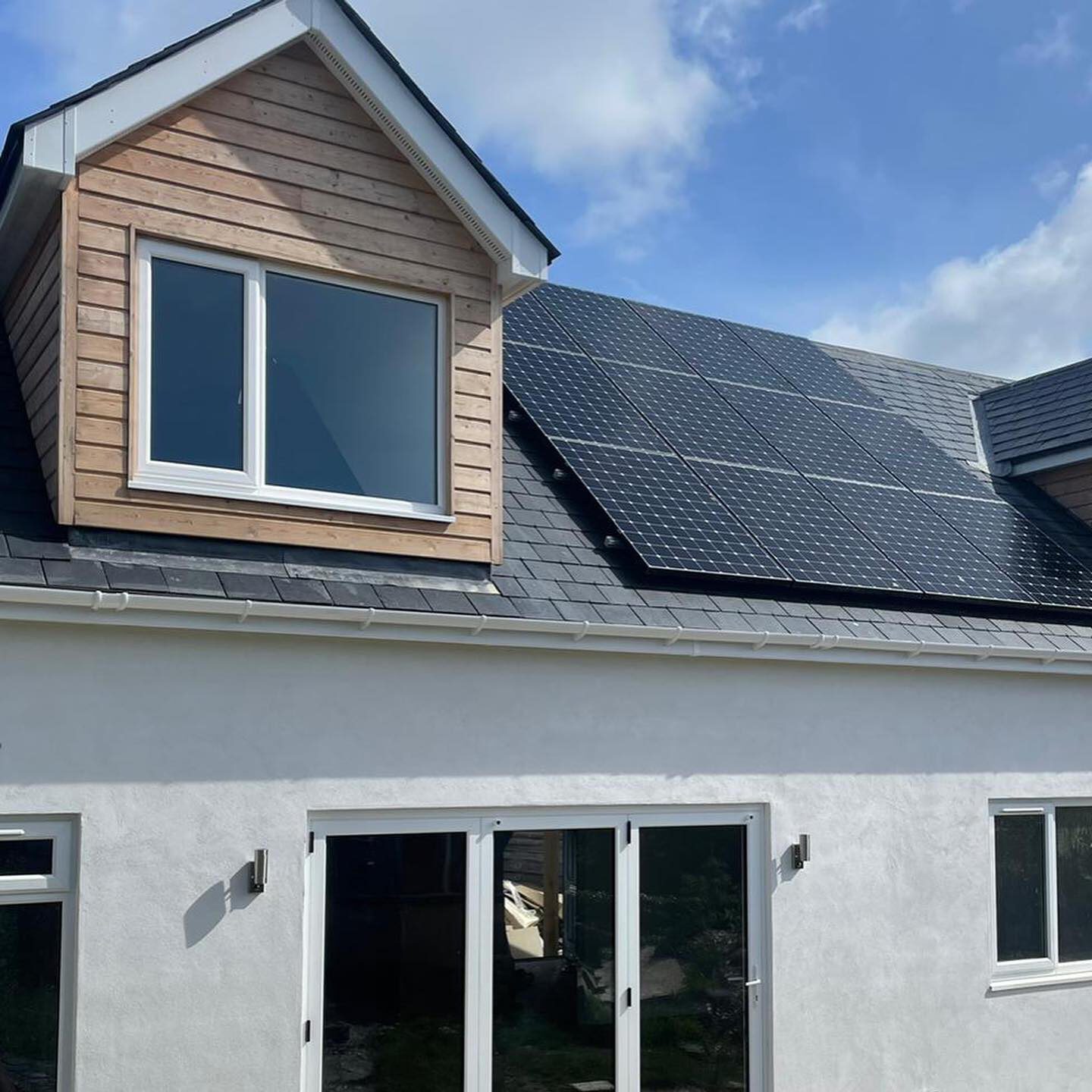 A white house with a dormer window, solar panels on the roof, and large glass patio doors. The ground outside appears unfinished, with some gravel and patches of grass. The sky is partly cloudy.