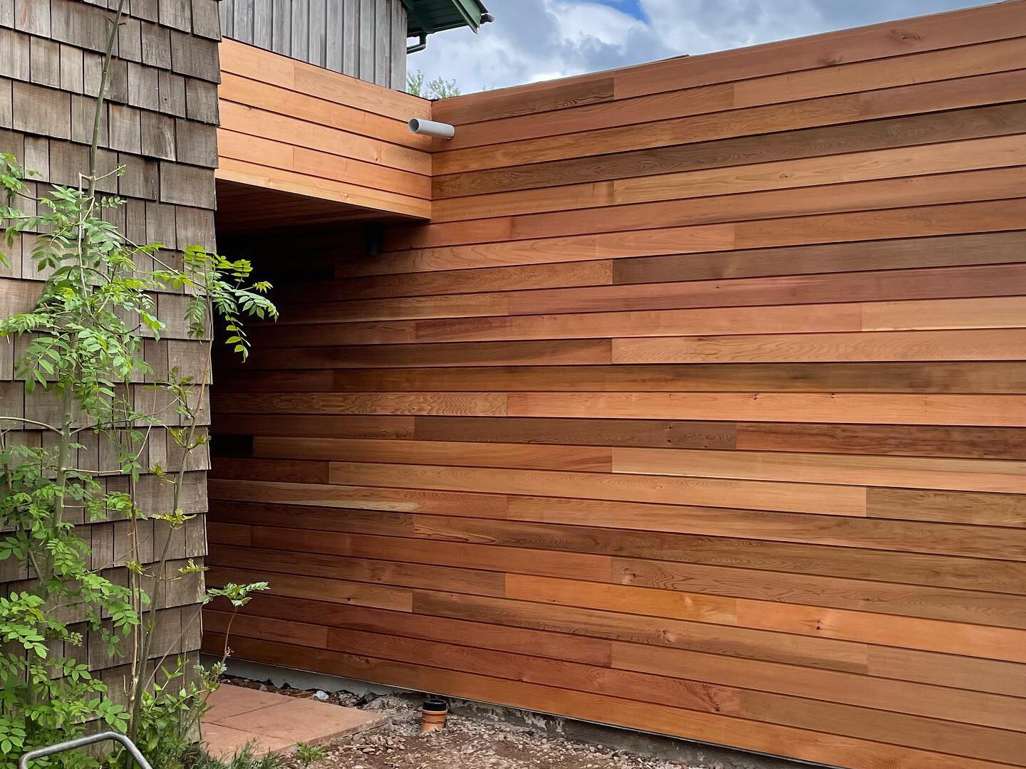 Outdoor view of a wooden house with horizontal planks and a shingled section. A cloudy sky is above, and a small plant grows near the corner. A power tool is visible on the ground in the foreground.
